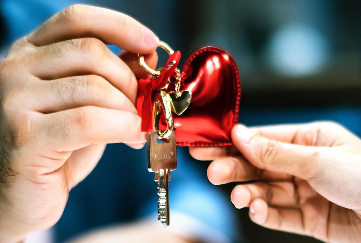 Two hands exchanging a key with a red heart-shaped keychain, symbolizing love, care, or a new home. The close-up image focuses on the key and keychain with soft lighting in the background.