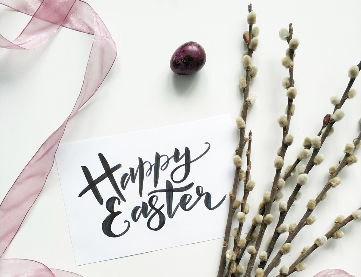 Flat lay of an Easter greeting with 'Happy Easter' written on a white card, accompanied by a soft pink ribbon, a purple speckled egg, and pussy willow branches on a white background