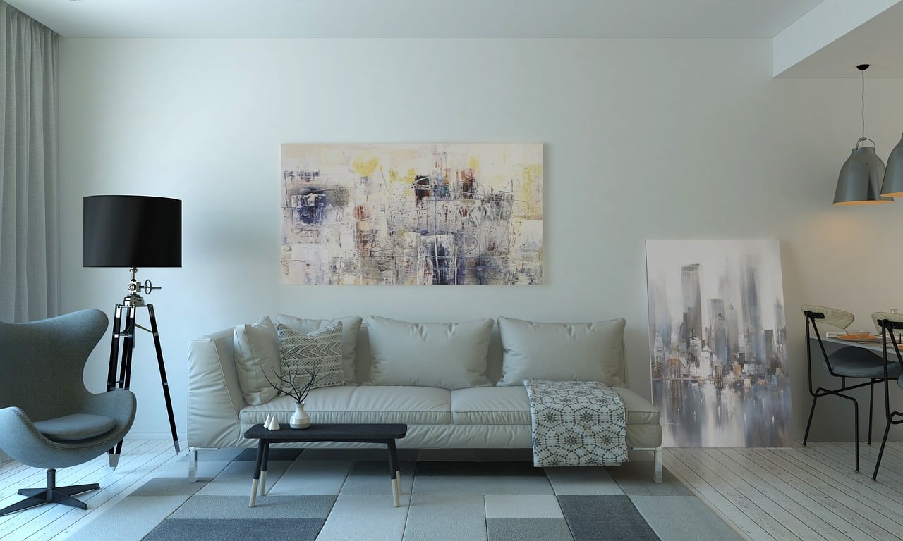 Modern living room with a grey sofa, abstract wall art, black floor lamp, and contemporary decor. The room has a neutral color palette with a grey accent chair, patterned rug, and a dining area in the background
