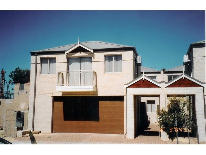 A contemporary double-story townhouse featuring a large brown double garage door, a balcony with metal railings, and light-coloured brick walls under a clear blue sky.