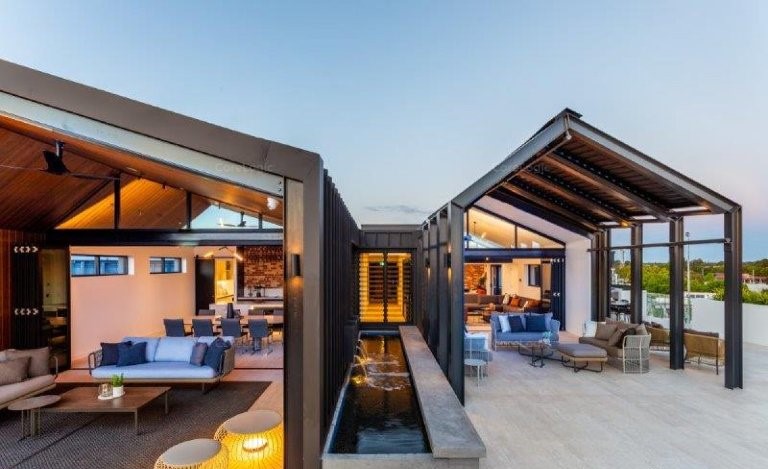 A modern luxury outdoor living area featuring open roof structures, cozy seating, and a sleek water feature.