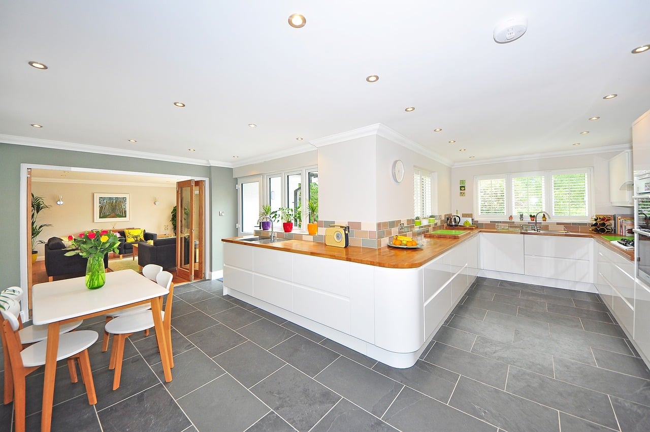 Spacious modern kitchen with white cabinets, wooden countertops, and a dining area with a small table and chairs. The kitchen features recessed lighting, tiled flooring, and large windows providing ample natural light.