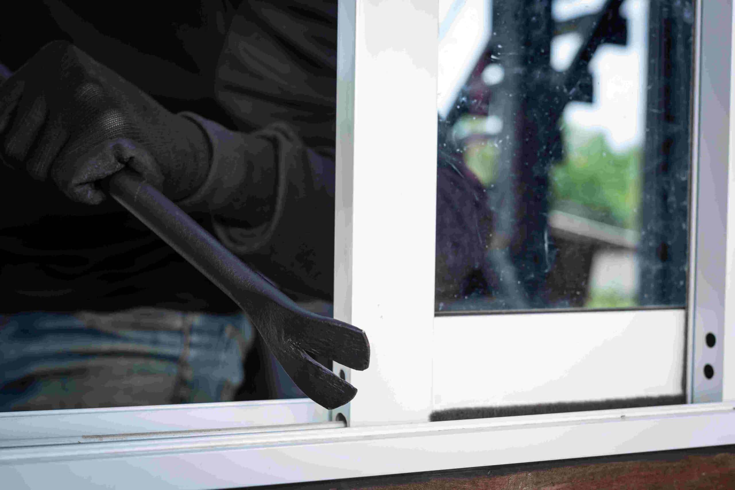 A close-up of a person in dark clothing attempting to pry open a window with a crowbar, highlighting the importance of home security measures.